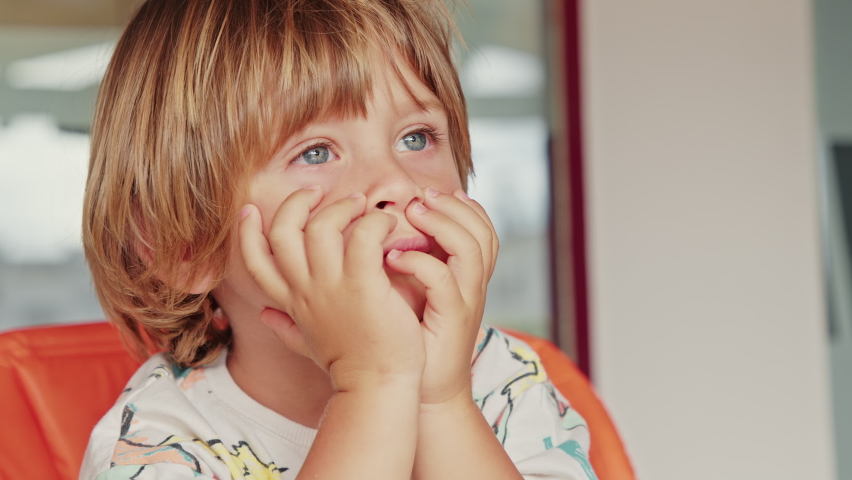 Portrait of a little lovely kid sitting in orange-colored chair in the kitchen. Caucasian, fair-haired boy playing alone indoors on a sunny morning. High quality 4k footage