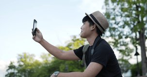 Side view Portrait of Happy Young Asian traveler man with hat video call on mobile phone while sitting on stairs at the park. Smiling Male speaking on cellphone at park. Hobby and Lifestyle concept.