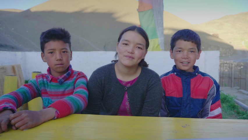 Shot of three young mixed-gender east Asian kids with smiles on their faces sitting outdoors together on a bench and looking at the camera in a mountainous cold region or terrain. Royalty-Free Stock Footage #1081552679