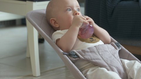 Cute baby boy drinking in the babychair at home, toddler sucking fruit juice from a baby bottle while sitting in rocking chair. High quality 4k footage