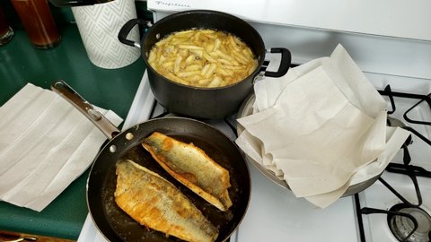 Home cooking - Two fillets of Walleye or Yellow Pike with skin being cooked on non stick pan while French fries being deep fried in non stick pot.