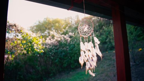 A beautiful cinematic shot of a knitted dreamcatcher with feathers and stones hangs on the veranda against the backdrop of the sun and a garden.