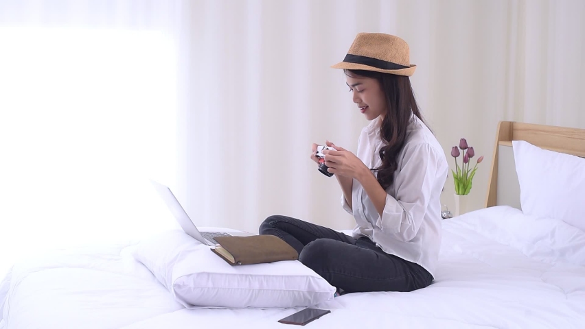Young Asian woman sitting on the bed and preparing for trip, using a laptop computer to searching information for travel is opening a country after quarantine in hotel during the coronavirus outbreak. Royalty-Free Stock Footage #1081558586
