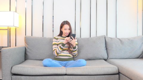 Asian woman play mobile games sitting on sofa at home and exult for victory rising fist