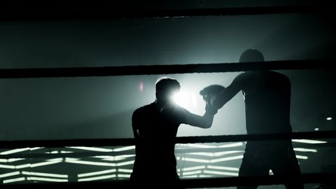 Boxer training and practicing with his trainer in low light gym . Silhouette on dark background. Boxer training in ring . Cinematic slow motion shot of professional young man punching with instructor