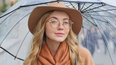 Close-up beautiful young girl walking on street in city with transparent umbrella portrait caucasian pensive woman with hat and glasses millennial lady looking distance enjoying rainy weather outdoors