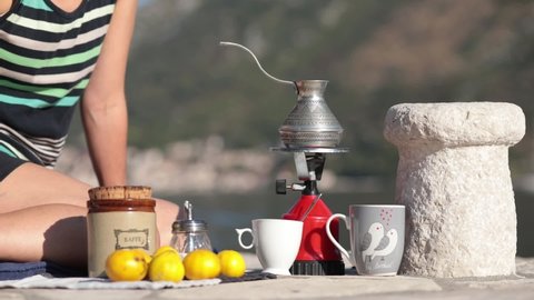 Unrecognizable woman sits on ground at picnic in Montenegro and make coffee in cezve on camp gas burner at pier in bay of Donji Stoliv. Woman make coffee outdoors with mountain blurred background