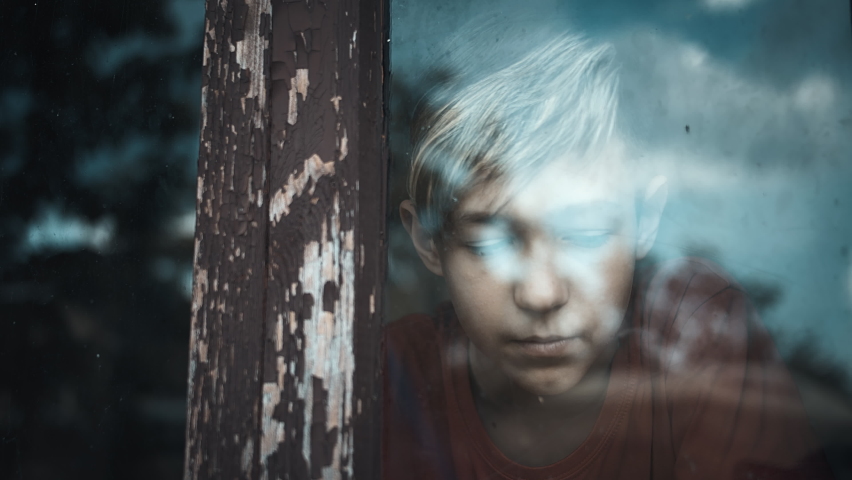 serious sad boy looks out the old shabby window and thinking about something, cinematic shot