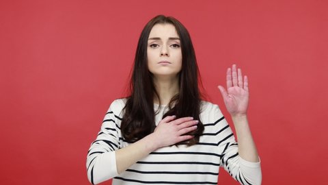 Young woman raising hand to take oath, promising to be honest, telling truth, pledging allegiance, wearing casual style long sleeve shirt. Indoor studio shot isolated on red background.