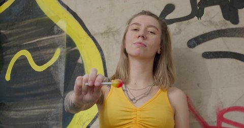 September 06 2021, St. Petersburg, Russia: Cheeky troubled teenage girl with tattoos on hands shows lollipop, tastes it and drops it, leaning against a concrete wall covered with graffiti, front view.