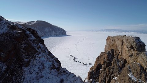 Aerial orbital shot of cape Khoboy, Olkhon island. Tall rocks in frozen lake Baikal with many people and cars around. Popular touristic destination. Winter landscape. Panoramic view