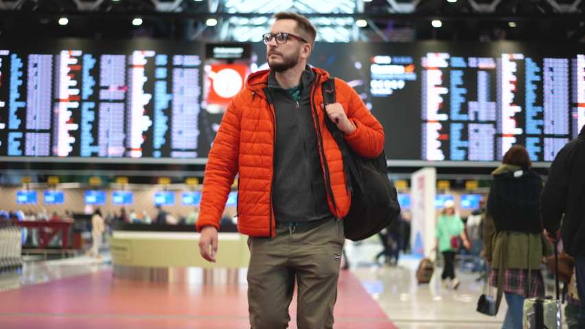 Travelling on vacation passenger person in glasses wearing orange jacket with backpack walking at airport terminal. People before arrival on runway. Flights check technology, modern airport board sign Royalty-Free Stock Footage #1081583042