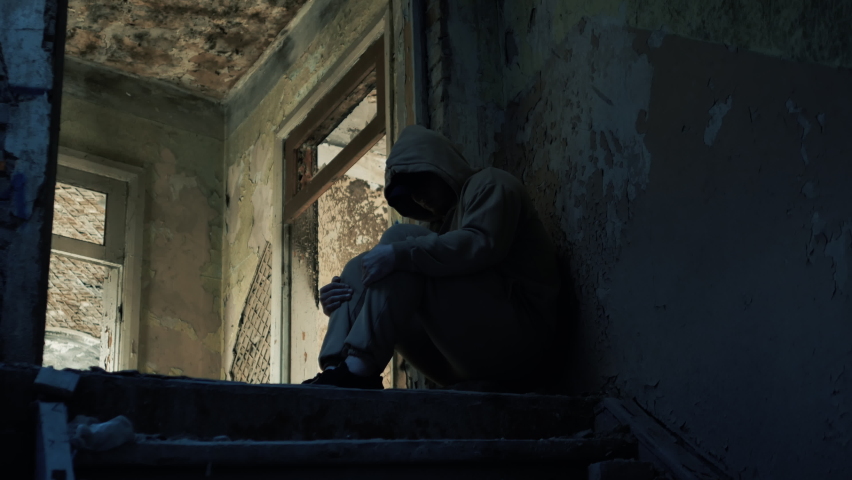 A young guy on the steps of an abandoned house in depression, a sad man, a person has social problems. The boy is wearing a hoodie in sadness. Earthquake destruction of the house.
