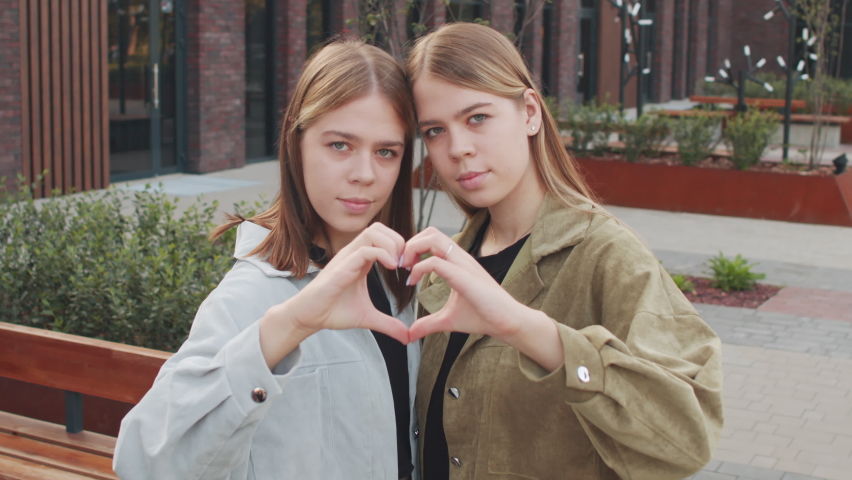 Tracking portrait of 20-year-old twin sisters doing hand heart gesture and posing for camera outdoors | Shutterstock HD Video #1081587551