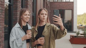 Tracking shot of cheerful young twin sisters holding coffee in to-go cups and recording vlog on their mobile phone outside - Powered by Shutterstock - Get 15% off with code: PIKWIZARD15