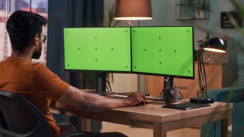 Bearded Indian guy browsing computers with chromakey monitors while sitting at table in dim room at home