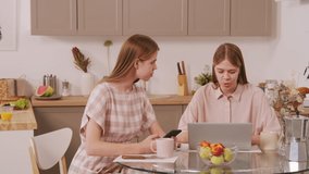 PAN shot of cheerful young twin sisters sitting at table in cozy kitchen and using laptop and mobile phone while chatting and enjoying their morning coffee - Powered by Shutterstock - Get 15% off with code: PIKWIZARD15