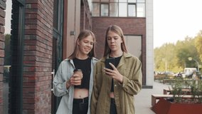 Tracking shot of cheerful young twin sisters walking down street and chatting while looking at mobile phone - Powered by Shutterstock - Get 15% off with code: PIKWIZARD15