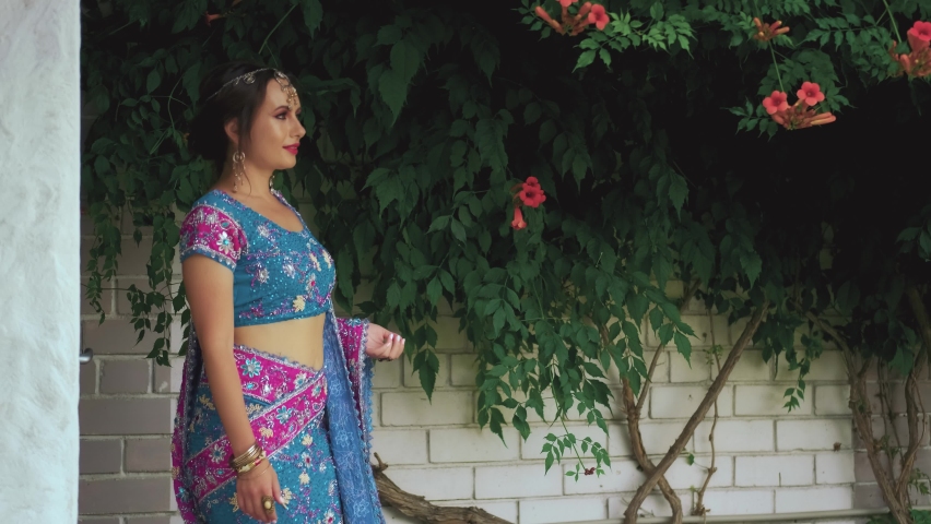 Oriental woman walks towards flowering shrub Kampsis. Indian adult girl princess enjoy scent natural flower. Blue pink traditional vintage dress clothing. Silver tiara. National bracelets adorn hand.