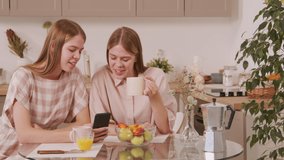 PAN shot of happy young twin sisters sitting at table in cozy kitchen and chatting while having coffee and browsing social media on mobile phone - Powered by Shutterstock - Get 15% off with code: PIKWIZARD15