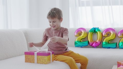 The Season of Giving concept. Happy smiling kid opens Christmas gift box near 2022 colorful balloons at home. Winter time, New Year presents and seasonal tradition - gift-giving.
