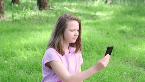 Teenage girl with smartphone having video call. Girl sitting on the lawn in city park and talking with relatives on cell phone