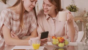 Tilt up shot of cheerful twin sisters sitting at table in cozy kitchen and chatting while looking at mobile phone and having coffee in morning - Powered by Shutterstock - Get 15% off with code: PIKWIZARD15