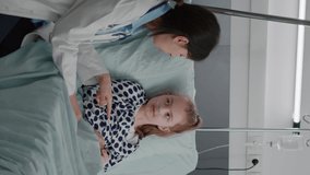 vertical video: Pediatric woman doctor interacting with sick patient giving high five during clinical consultation in hospital ward. Child lying in bed wearing oxygen nasal tube waiting for medication
