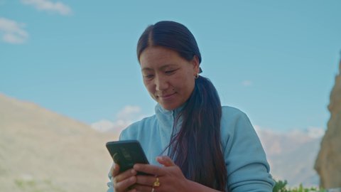 close shot of an Asian adult female solo traveler or backpacker standing outdoors in the high altitude region and using a mobile phone or smartphone with the mountains in the background 