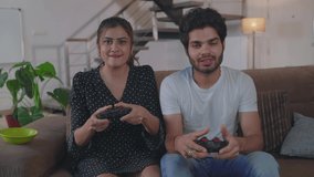 A young Asian Indian happy and cheerful modern couple sitting in front of a screen on a couch and playing a video game holding joysticks in their hands in an interior home setup 