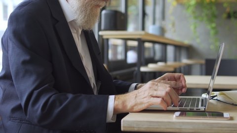 Senior teacher with grey beard typing on laptop, sitting at table, technology