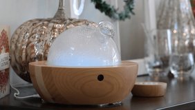 An essential oils diffuser that is made of wood and glass. It changes color and produces a steady stream of mist. There's a remote next to it and it's all sitting on a wooden table in a living room.