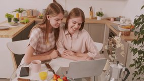 Handheld shot of happy twin sisters wearing pastel colored clothes sitting at table in cozy kitchen and laughing while chatting and using laptop