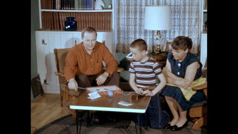 1950s: Man and woman sitting in chairs help a boy stretch out a wire attached to a toy telegraph key. Slate. Boy takes off headdress then starts assembling a toy telegraph. He read instructions.