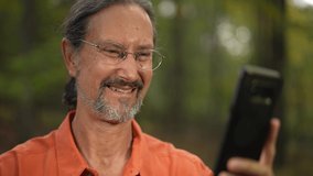 Portrait of ethnic, mature man smiling and waving to someone on video chat on smartphone in a natural outdoor environment.
