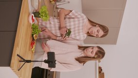 Vertical shot of identical twin sisters cooking in kitchen and recording video on mobile phone