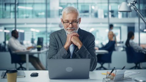 Modern Office: Portrait of Successful Middle Aged Bearded Businessman Working on a Laptop at his Desk. Smiling Corporate Worker. Multi-Ethnic Workplace with Happy Professionals. Front View Static Shot