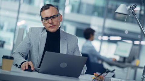 Modern Office: Portrait of Successful Middle Aged Businessman Wearing Suit and Glasses Working on a Laptop at his Desk. Serious Confident Corporate Executive. Front View Static Dutch Shot