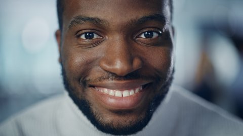 Close-up Portrait of Handsome Black Man with Deep Brown Eyes, Trimmed Beard, Wearing Stylish White Turtleneck, Winks and Shows Perfect Smile. Attractive Authentic Gentleman Turns, Looks at the Camera