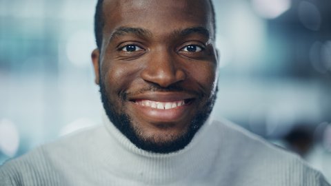 Close-up Portrait of Handsome Black Man with Deep Brown Eyes, Trimmed Beard, Wearing Stylish Turtleneck, Smiles Charmingly. Attractive Authentic African American Gentleman Looks up at the Camera