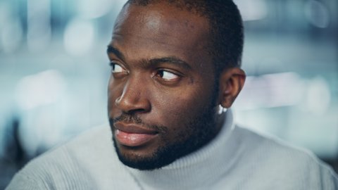 Close-up Portrait of Handsome Black Man with Deep Brown Eyes, Trimmed Beard, Wearing Stylish White Turtleneck, Winks and Shows Perfect Smile. Attractive Authentic Gentleman Turns, Looks at the Camera