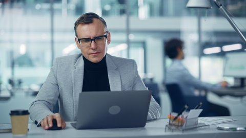 Modern Office: Portrait of Successful Middle Aged Businessman Wearing Suit and Glasses Working on a Laptop at his Desk. Serious, Confident, Stylish Corporate Executive Using Computer. Front View Shot
