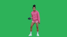 Portrait of young woman table tennis player over green background. African american girl wearing pink hoodie and shorts having fun with racket. Chroma key. Copy space. 4k raw video footage