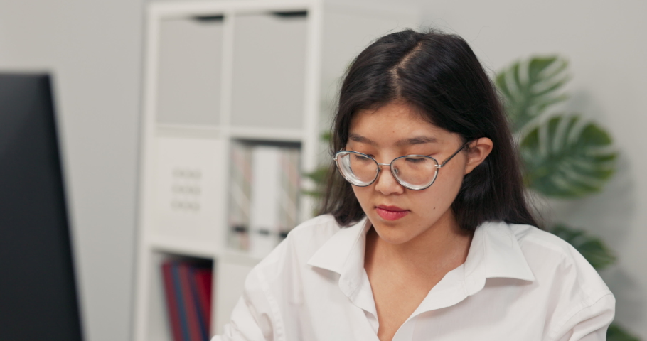 Beautiful secretary in glasses with characteristic Korean Asian beauty dressed in white shirt, sits in the office in front of the computer, working, in the background co-workers walk around company Royalty-Free Stock Footage #1081615607