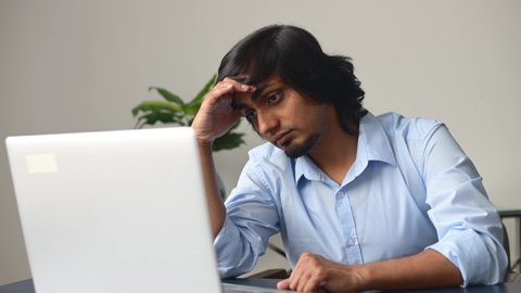 Upset young indian freelance guy sitting at the desk, using laptop, feels tired and exhausted, sad eastern man worrying, feels anxiety about problems, alarming