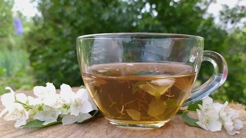 Cup of green tea with flowers jasmine. Jasmine tea and jasmine flowers on wooden background. Jasmine Flower and Leaf. Slow motion pour tea in a cup and hand teaspoon stir close up.