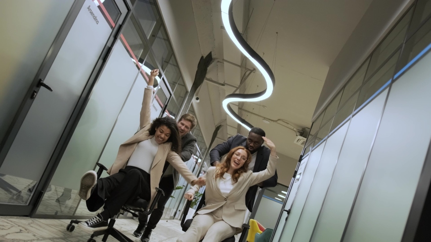 Excited young cheerful multiracial diverse colleagues having fun riding on office chairs in coworking space. Active happy millennial workers take a break enjoy play game laughing together at workplace | Shutterstock HD Video #1081621187