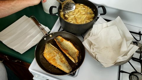 Home cooking - Two fillets of Walleye or Yellow Pike with skin being cooked on non stick pan while removing French fries from pot into bowl with paper.