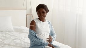 Vaccinated Black Pregnant Woman Looking At Her Arm With Adhesive Bandage After Vaccination And Tenderly Touching Belly While Sitting On Bed At Home, Got Injection Shot Against Covid-19, Slow Motion