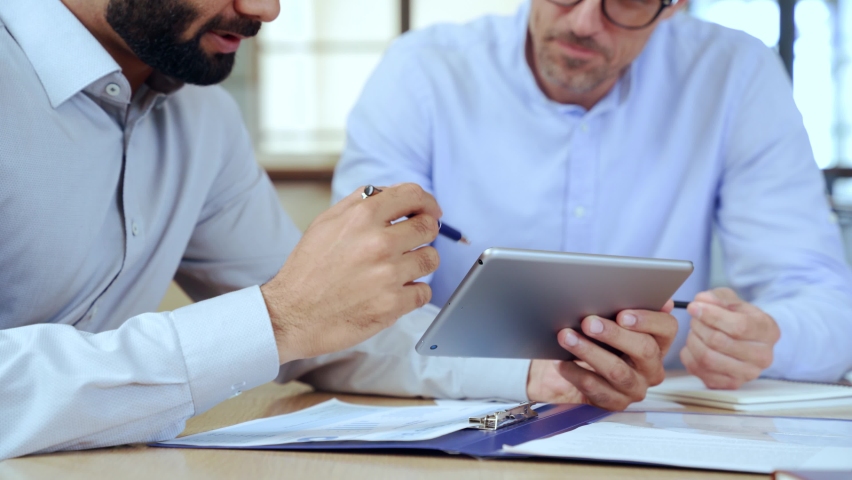 Two diverse business men discussing analyzing financial market data using digital tablet. Financial advisor broker manager consulting investor client managing project tech at office meeting. Closeup Royalty-Free Stock Footage #1081628171